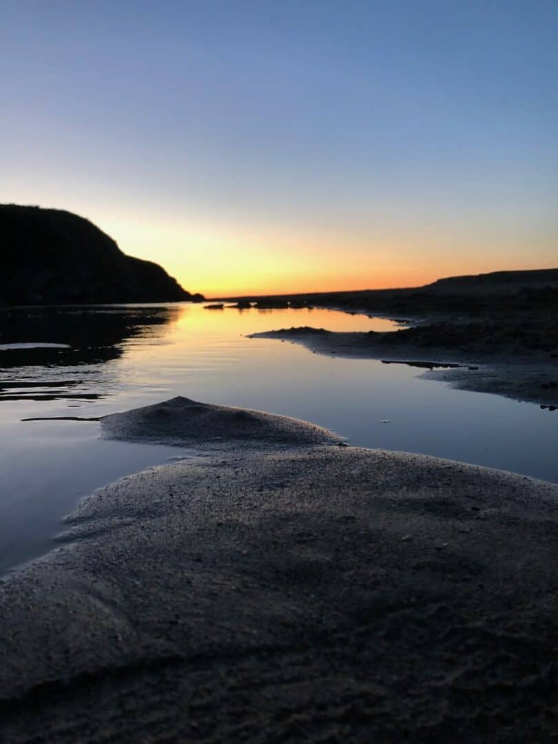 An image of the sunset hitting the surface of a body of water. The sun silhouettes stones and hills in the image. 