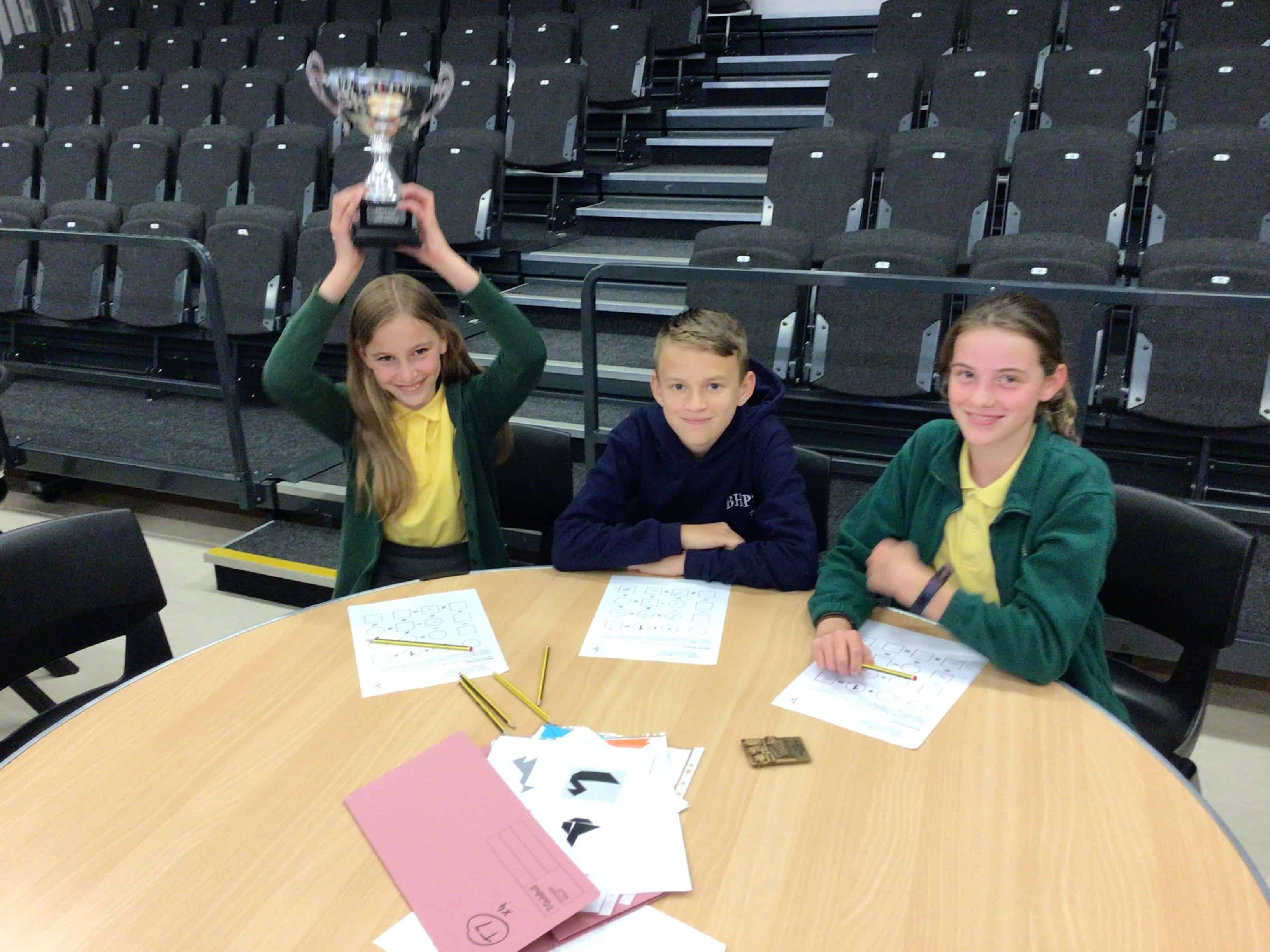 The 3 winning pupils from Bradshaw Hall Primary smile at a table, one of them holds the CHHS Maths Challenge trophy