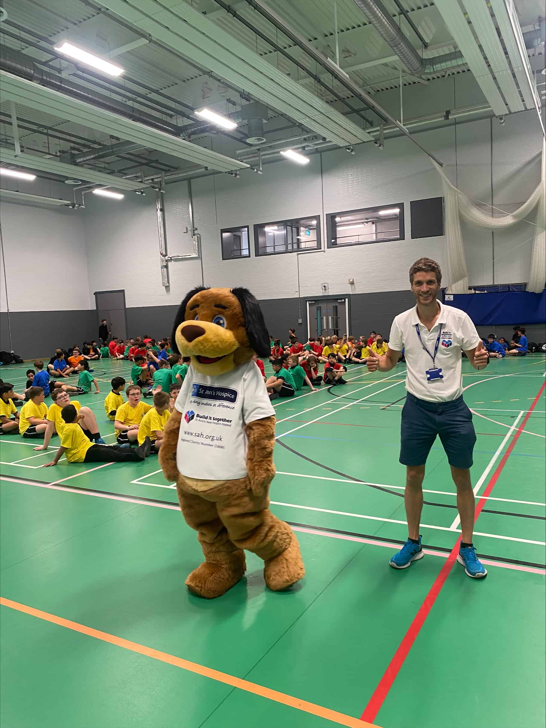 Year 7 students from Cheadle Hulme High School sit in the sports hall wearing their House team shirts, Stan the St Ann's Hospice mascot dog stands next to a teacher in front of the crowd of students.