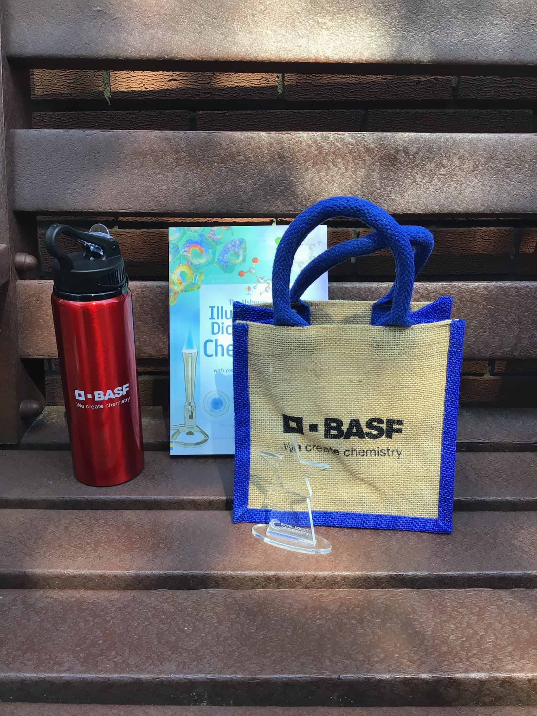 The items received by the Cheadle Hulme High School students who competed in the Chemquiz. These are: A BASF red water bottle, a book about Chemistry, a BASF tote bag, and a Royal Society of Chemistry trophy.