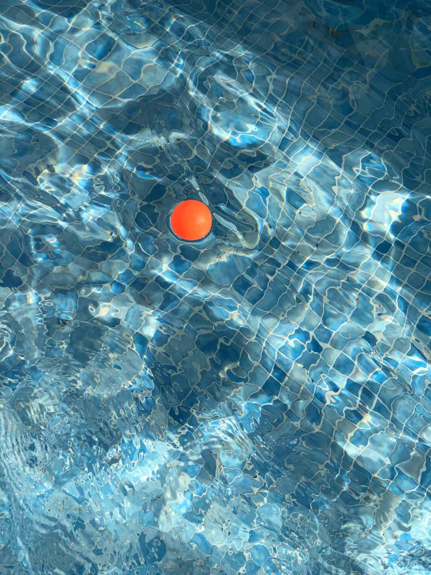 A bright orange ping-pong ball contrasts the light blue water as it floats in a swimming pool.