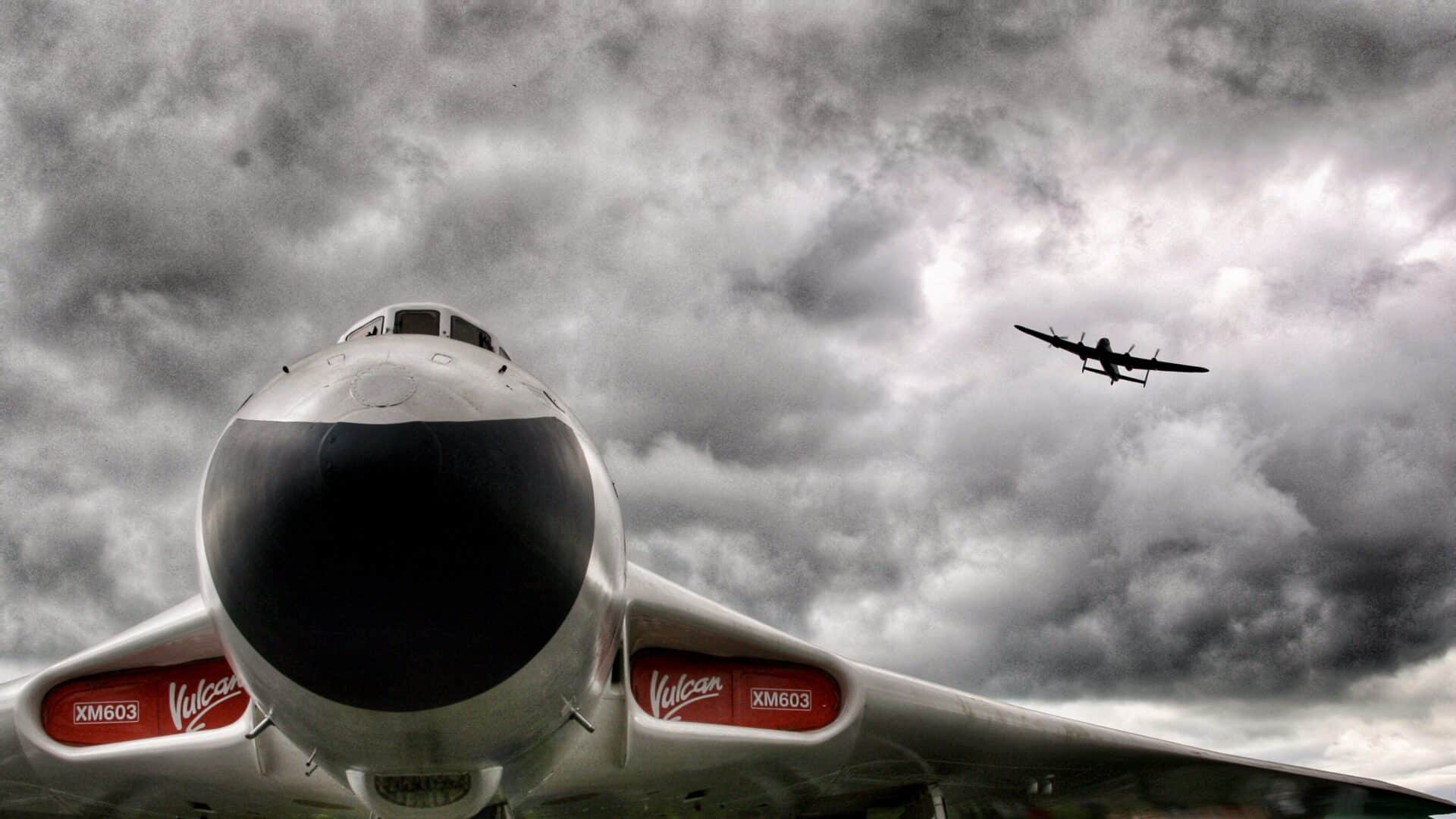 The nose of an airplane foregrounds this shot with its wings spanning the lower third of the image. A plane can be seen in flight in the distant background. The background is made up of grey storm clouds.