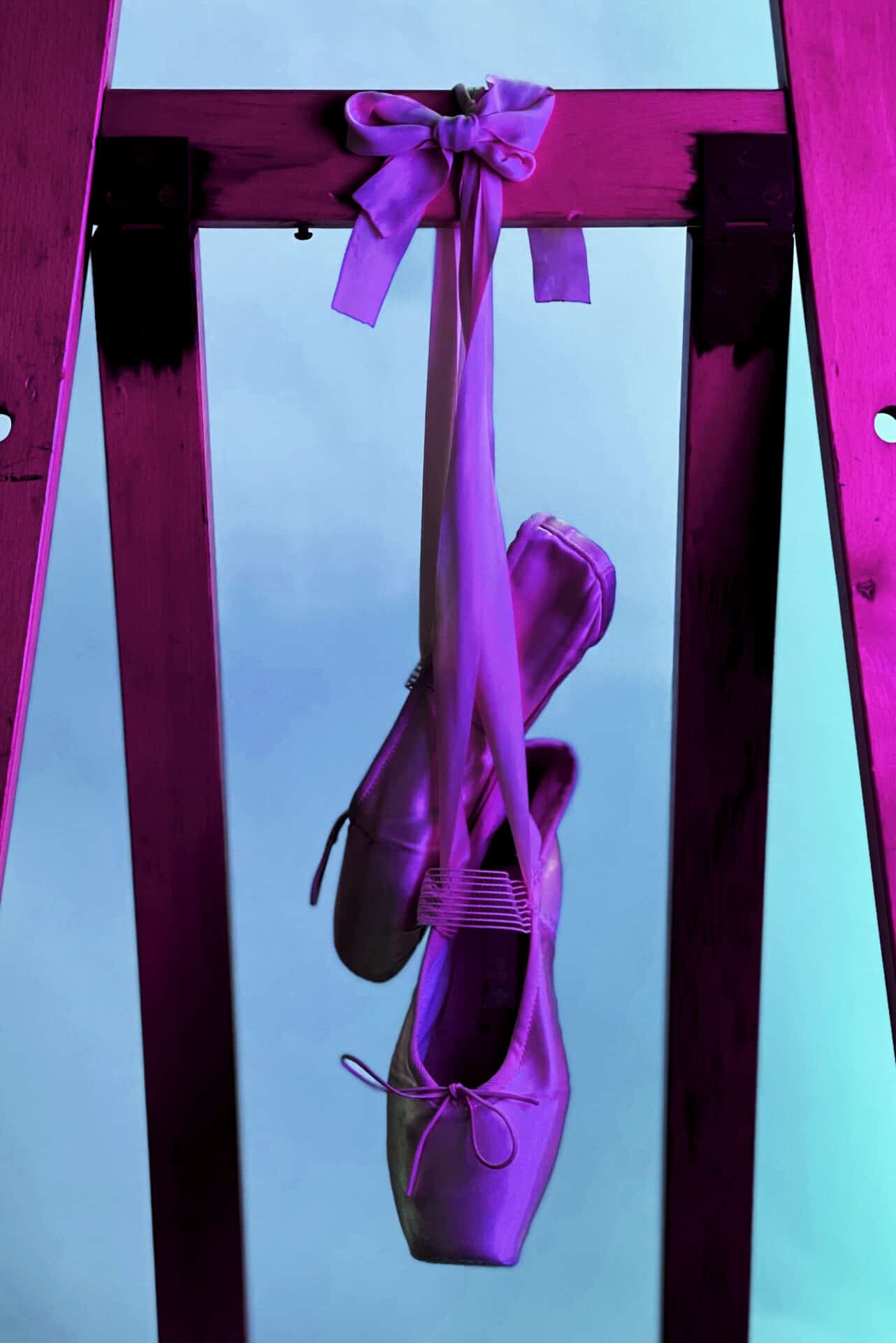 A close-up of ballet shoes hanging from a bar in a dance studio. The image is full of blue, purple, and pink tones that appear to come from a light source within the room.