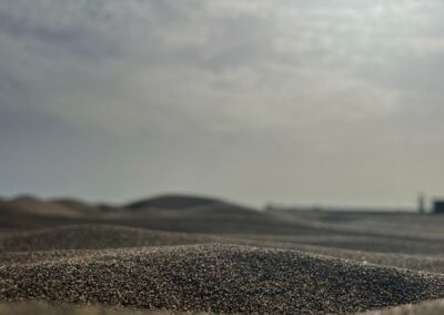 A close-up on a mound of dark sand on a beach. The background is out of focus but full of clouds. The sun's light creates contrasting tones in the image.